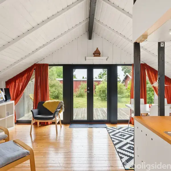 Et lyst loftsrum med skråt loft og træmøbler, har store glasdøre, der åbner ud til en have. Røde gardiner hænger ved siden af dørene, og et sort/hvidt tæppe ligger på gulvet.