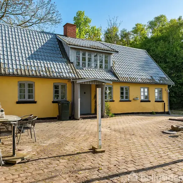 Et gult hus med gråt tag står i en gårdsplads omgivet af træer. Der er et udendørs bord og stole til venstre, og en postkasse ved hoveddøren.