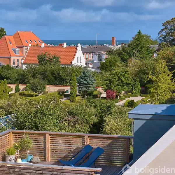 Terrasse med træmøbler og planter i et frodigt haveområde. Baghuse med røde tage ses i baggrunden, sammen med havudsigten under en skyet himmel.