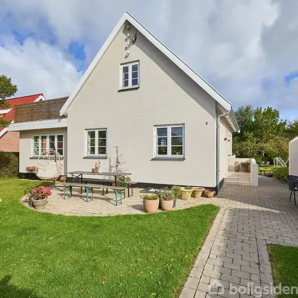 En hvid, to-etagers hus står i en velplejet have med grøn græsplæne. Et flisebelagt område har et udendørs bord med stole. Blå himmel med spredte skyer i baggrunden.