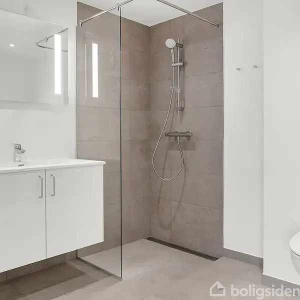Et moderne badeværelse viser en bruser med glasvæg, hængende toilet og hvidt badeværelsesskab med vask. Væggene er lyse, og der er indbygget lys ved spejlet.