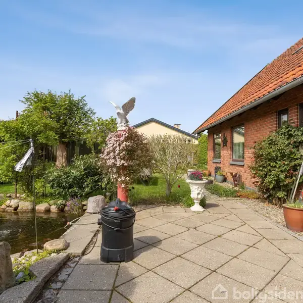 En skulptur med fugl står på en terrasse i en have ved et murstenshus. Der er en dam med sten, planter og en anden dekorativ skulptur.