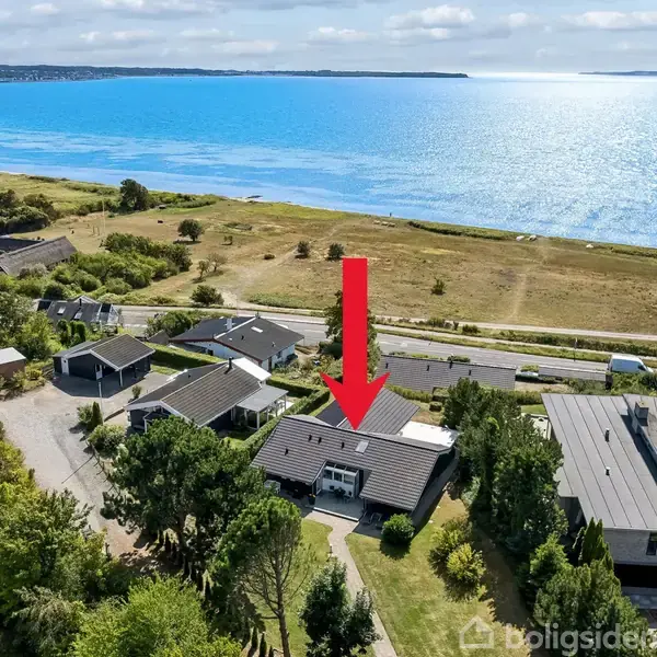 Hus markeret med en rød pil, placeret blandt andre huse, tæt på kystlinjen med udsigt over havet under en solrig himmel.