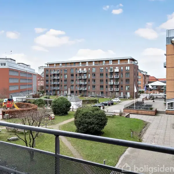 Bygning ses fra en altan i et gårdmiljø. Foran byggeriet er der en græsplæne, gangstier og nogle buske. I baggrunden står flere etagebygninger under en blå himmel med skyer.
