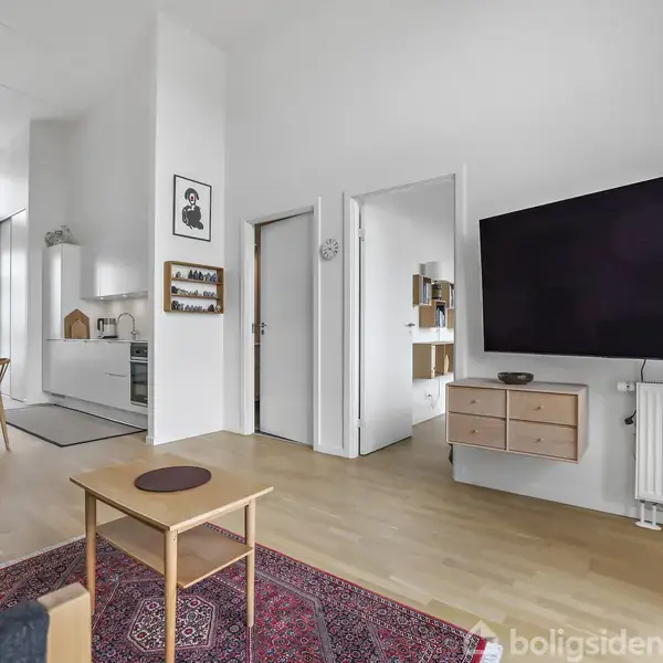 Rummet indeholder en stue med en stor fladskærm på væggen, en sofa og et sofabord på et tæppe. Til venstre er et åbent køkken- og spiseområde.