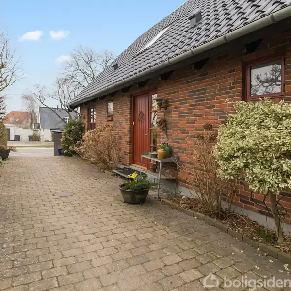En muret stibet villa med en rød dør og vinduer, flankeret af planter og buske. Indkørsel med brosten fører op til huset, omgivet af en rolig, beboet gade.