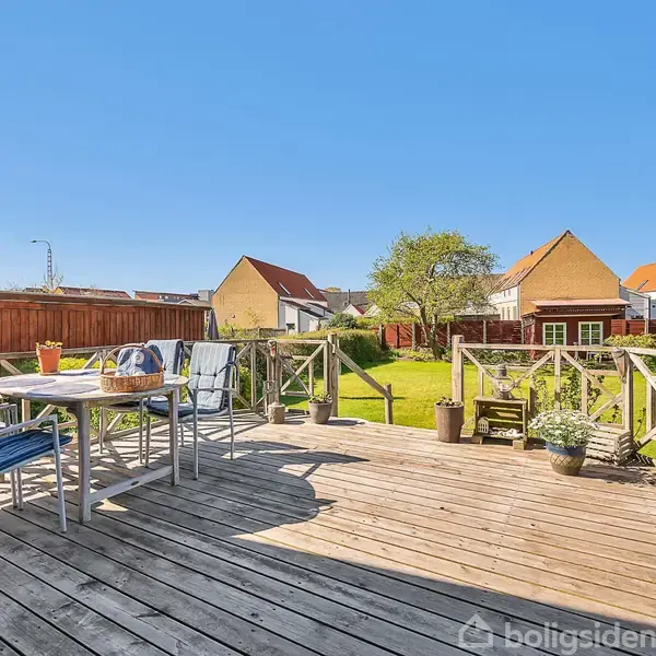 Træterrasse med bord og stole, badet i sollys. Omgivet af grøn have med buske og huse i baggrunden under blå himmel. Rolig og idyllisk atmosfære.