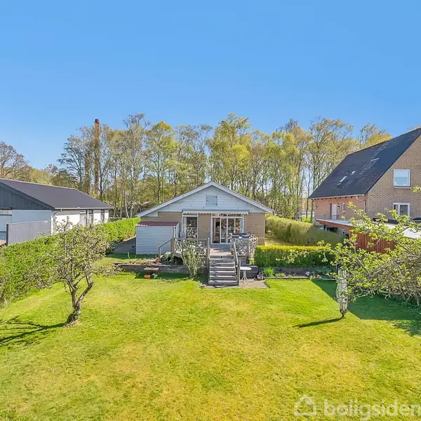En charmerende hvid bungalow med en anlagt have og træterrasser. Huset er omgivet af grønne hegn og ligger i et skovområde med andre moderne boliger i nærheden.