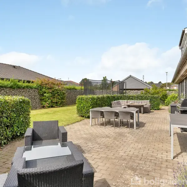 Terrasse med sorte kurvemøbler og borde med stole står på flisebelagt område. Omkranset af hække og græsplæne. Hus med sort træfacade i højre side under blå himmel.