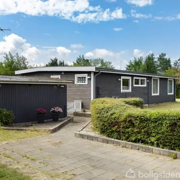 Gråt sommerhus står stille ved siden af en garage. Omgivet af en trimmed hæk og flisebelagt gangsti under en blå himmel med skyer.