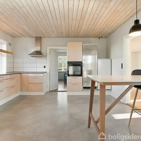 Et lyst køkken med træloft og flisegulv inkluderer en komfur med emhætte, indbyggede træskabe, og moderne hvidevarer. Bordområde med sort lampe over. Sollys strømmer ind gennem store vinduer.