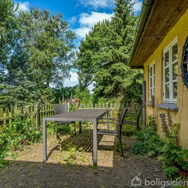 Have og terrassebord med stole står på en flisebelagt terrasse ved siden af et gult hus. Omkring er der et hegn og mange træer under en blå himmel.
