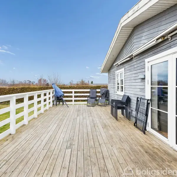 Træterrasse med stole står ved et gråt hus med store glasdøre; omgivet af en grøn græsplæne og hæk under en blå himmel.
