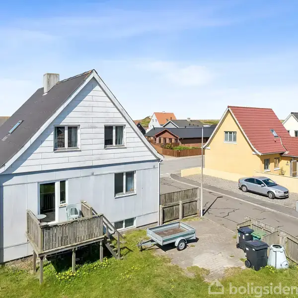 Hus med grå facade og skråtag står i en forstad. Foran huset er en lille terrasse og en trailer. Omkringliggende huse ses under en blå himmel.