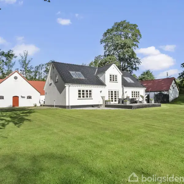 Et hvidt hus med sort tag står i en have med en velplejet græsplæne. Baghaven er omkranset af træer, og der er en terrasse med stole.