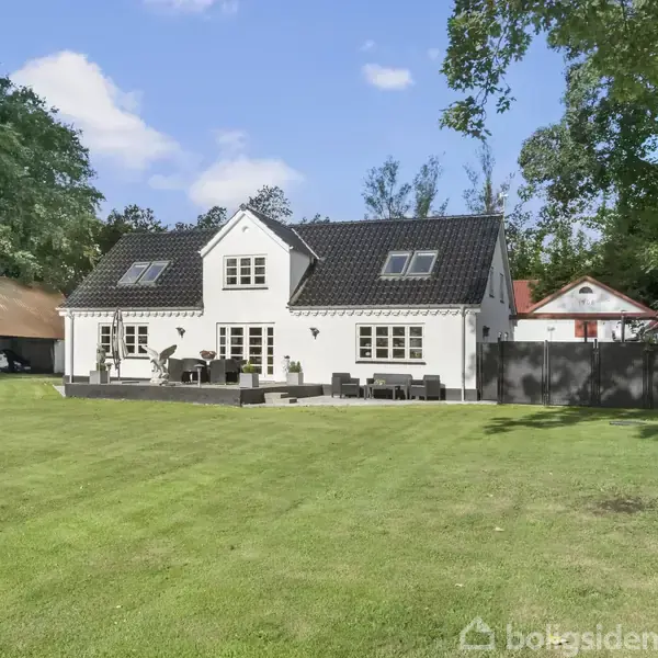 En hvid, to-etagers hus står stille omgivet af frodig græsplæne og tæt bevoksede træer. Terrassen foran huset har møbler, mens en carport ligger til venstre.