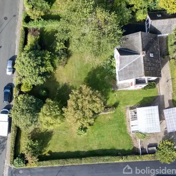 Hus set fra oven med have omgivet af træer og buske, en carport og terrasse. To veje løber parallelt til grunden med parkerede biler langs kanten.