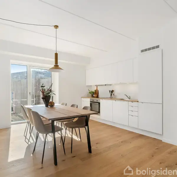 Et lyst køkken-alrum med et træbord omgivet af seks stole. En ovn og køkkenskabe er synlige til højre, med dekorationer på bordet og en lampe hængende over.