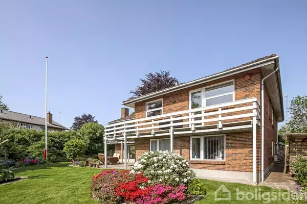 Et to-etagers rødstenshus med balkon står i en velplejet have med græsplæne, blomsterbede og flagstang. Solskin og træer omkranser huset.