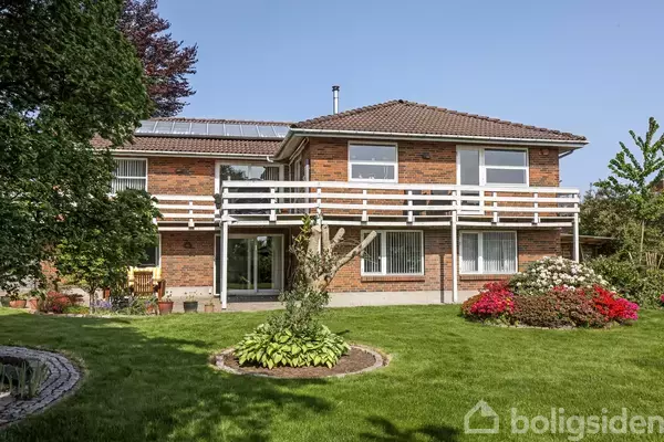 Et to-etagers hus med murstensfacade og balkon har vinduer med skodder. Det står i en velplejet have med grøn græsplæne, blomsterbede og træer, under en blå himmel.
