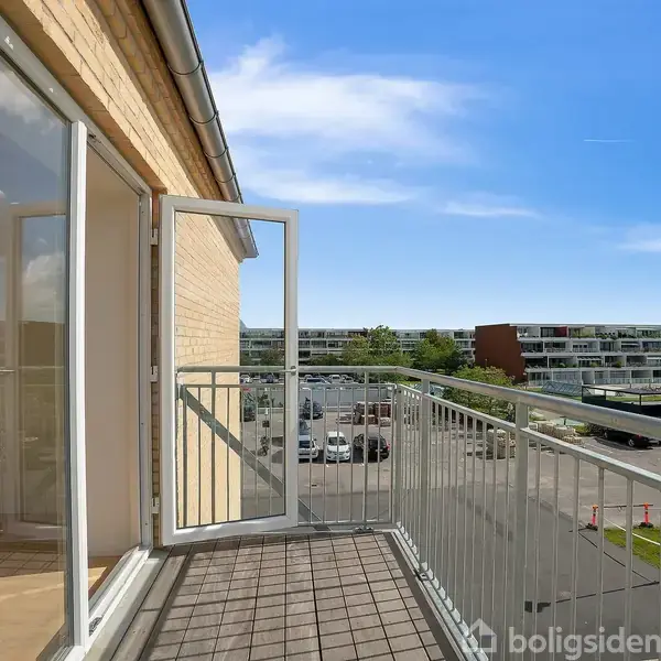 En altan med åben dør vender ud mod en parkeringsplads og lejlighedskompleks i baggrunden under en blå himmel.