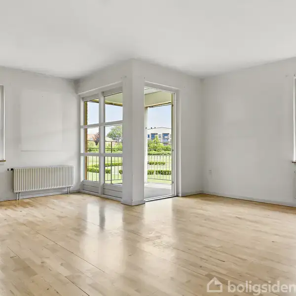 Et lyst rum med trægulv og to radiatorer. Store vinduer og en glasdør åbner ud til en balkon. Udsigt til grønne områder udenfor.