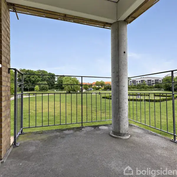 Balkon med jerngelænder omfatter en søjle og udsigt til en velplejet park med græsplæne, stier og træer. Moderne bygning er synlig i baggrunden under en blå himmel.