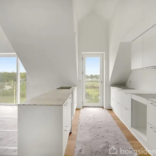 Et moderne, hvidt køkken med indbyggede skabe og skuffer er opstillet på begge sider af et smalt rum. To store vinduer giver udsigt til grønne områder udenfor.