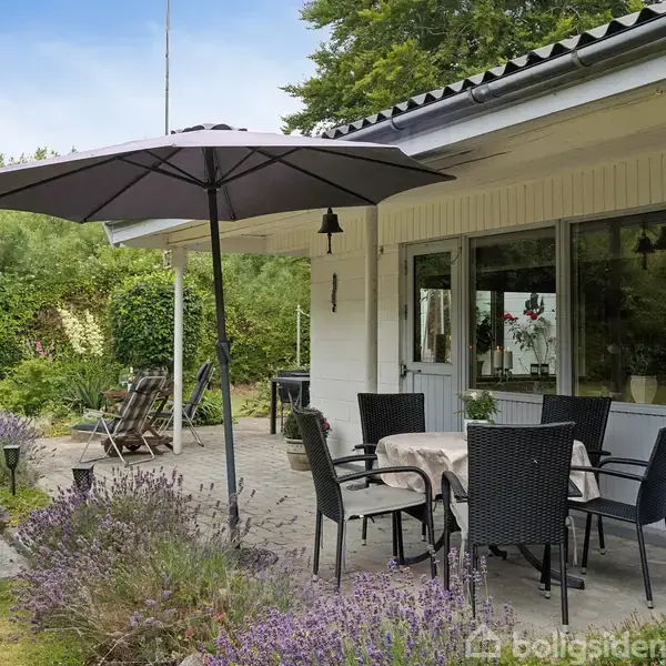 Terrassemøbler under en stor parasol står på en flisebelagt terrasse med lavendelbed ved siden af et hvidt hus. Et bord med dug og stole skaber en hyggelig udendørs spiseplads.