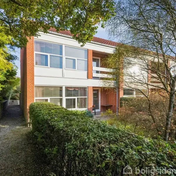 En to-etagers murstensbygning står omgivet af buske og træer, med en grussti til venstre, under en solfyldt og klar himmel.