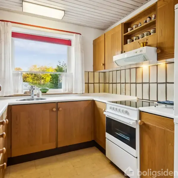 Køkken med træskabe og hvidevarer. Komfuret står ved hjørnet under en emhætte. Vinduet har rødt gardin og giver udsigt til træer. Vægge er beklædt med fliser.