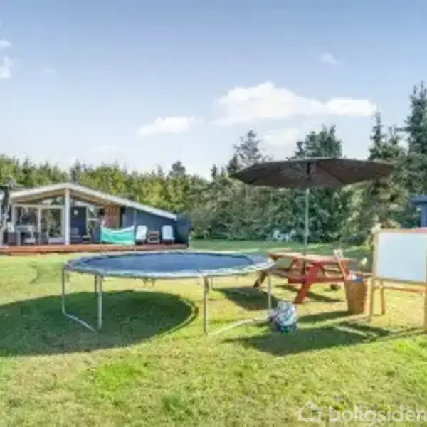 En trampolin står på en græsplæne foran et sommerhus. Ved siden af er et picnicbord med parasol og tavle. Omgivet af træer, skaber det en fredfyldt haveindstilling.