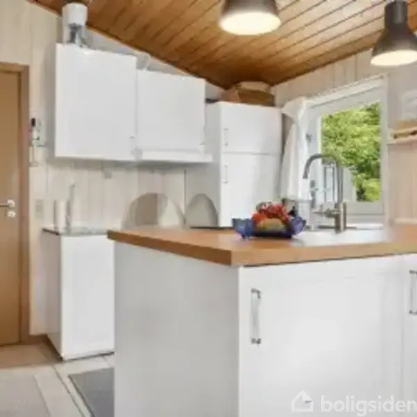 Køkken med hvidt skab og køleskab, en frugtskål på bordet under loftslamper. Trævægge og lofter. Skilt ved vinduet: "Happy is what happens when you love what you have."