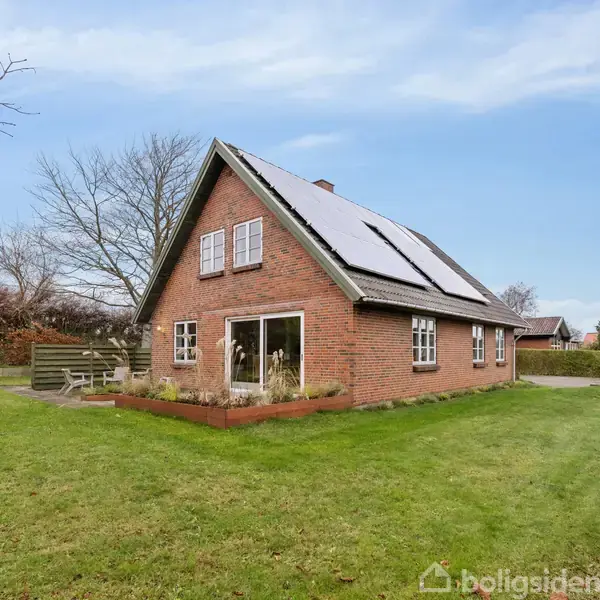 Et rødstenshus med solpaneler på taget står i en velholdt grøn have. Der er store vinduer og en terrasse, omgivet af træer og buske under en klar blå himmel.