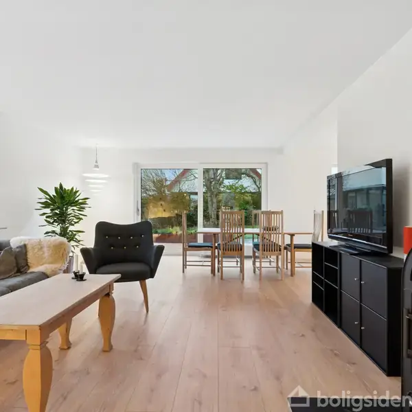 Stue med brændeovn står til højre, mens en sort skænk med fjernsyn ses foran. Sofa med sofabord til venstre, og i baggrunden er spisebord ved vindue med havudsigt.