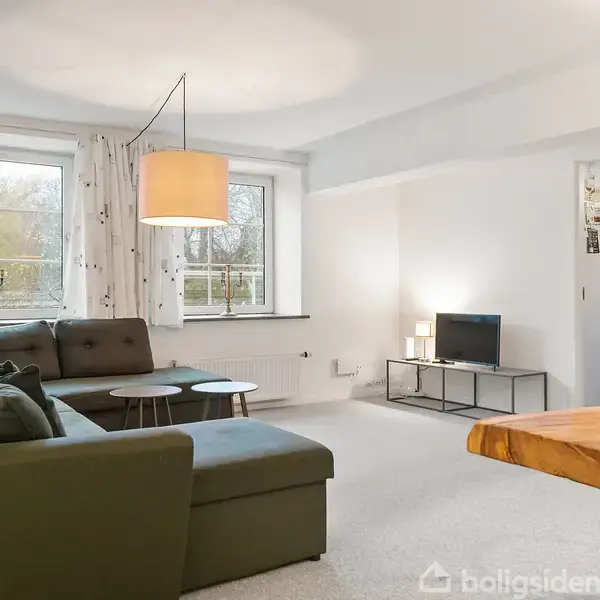 En stue med en mørkegrøn hjørnesofa placeret under to vinduer med hvide gardiner. Et rundt sofabord står foran sofaen. I baggrunden ses et fjernsyn og en åbning til et andet rum.