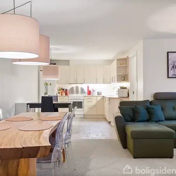 Træbord med dækservietter står ved siden af en grøn sofa i en lys stue med åbent køkken. Væglamper og billeder dekorerer rummet.