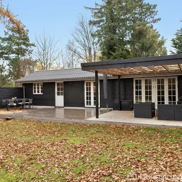 Sort træhus med en overdækket terrasse, omkranset af en stor have dækket med efterårsblade. Flere hvide døre og vinduer åbner ud til terrassen. Skovområde i baggrunden.