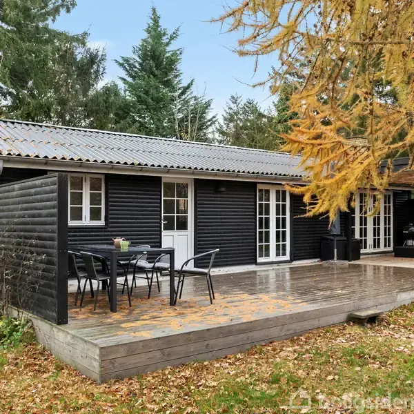 Et sort træsommerhus med hvidrammede døre står stille på en træterrasse med havemøbler. Omgivelserne inkluderer frodig vegetation og træer, der indikerer en skov- eller haveomgivelse.
