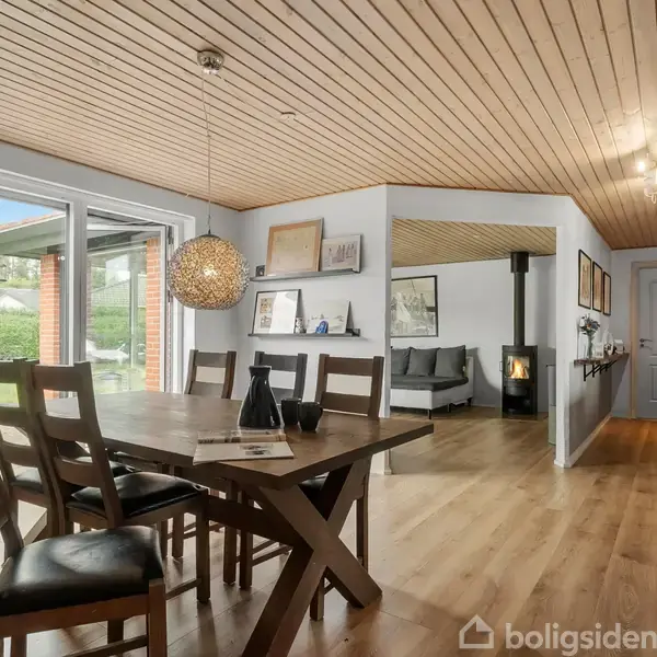 Spisebord med stole står i et lyst køkken-alrum. En rund lampe hænger over bordet. Vinduer viser en grøn have. En brændeovn er i det tilstødende opholdsrum.