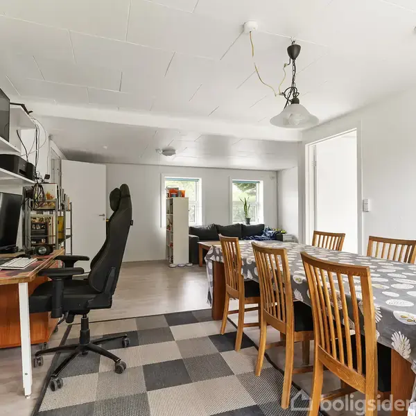 Kontorstol ved skrivebord med computer og kontorartikler; ved siden af står et spisebord med seks træstole og en dug med løvmønster i rum med hvide vægge og gulvlampe.