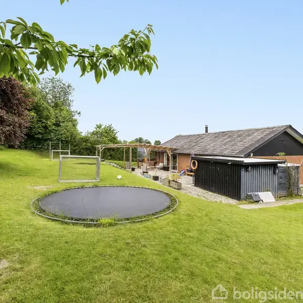 Hus ligger i en frodig have med græsplæne. Trampolin og gyngestativ findes i haven. Træer omkranser området, skabende en privat atmosfære.