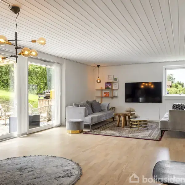 Stue med grå sofaer arrangeret rundt et sofabord på gulvtæppe. En fladskærm hænger på væggen. Store vinduer åbenbarer en terrasse med havemøbler og grøn have. Brændeovn i forgrunden.