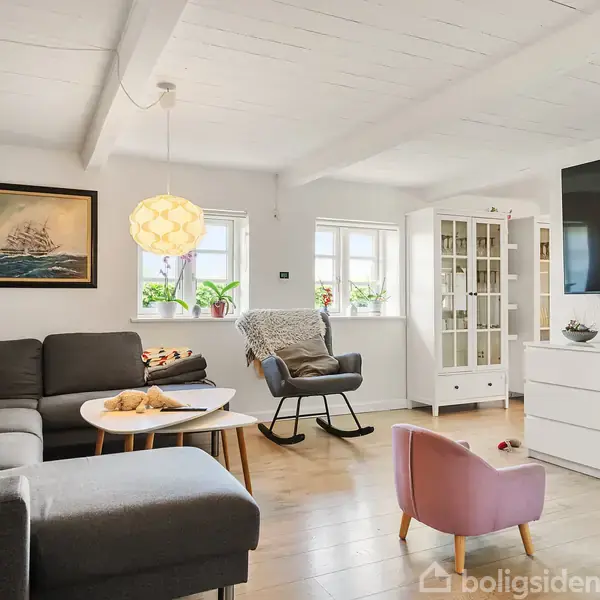 Stue med grå hjørnesofa, sofabord og gyngestol ved siden af et vindue. Stuen har træpaneler, fladskærms-tv og et rosa børnestol foran hvidt skab. Vægkunst over sofaen.