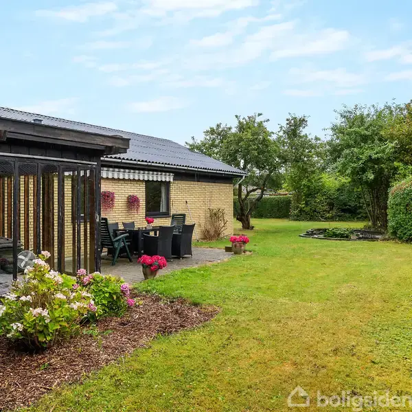 En gul murstensvilla med sort tag står ved siden af en velholdt have. Foran huset er der udendørs havemøbler og blomsterbede. Baggrunden har træer og grøn bevoksning.