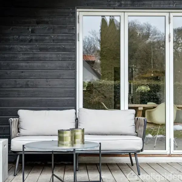 En udendørs sofa med lyse hynder står på en træterrasse foran en bygning med sorte træpaneler. Et lille bord står foran sofaen. Indendørs ses en spisegruppe gennem glasdøre.