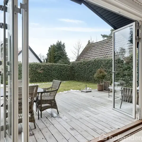 Folded glasdøre åbner ud til en træterrasse, hvor der står et bord med stole. I baggrunden ses en have med en grøn hæk og andre huse.