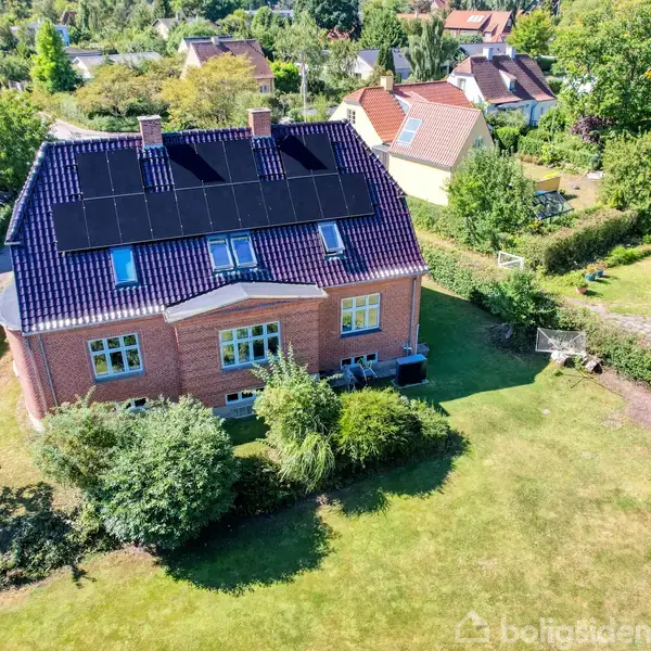Et rødt murstenshus med solpaneler på taget står i en grøn have. Omkring huset ses en række træer og buske.