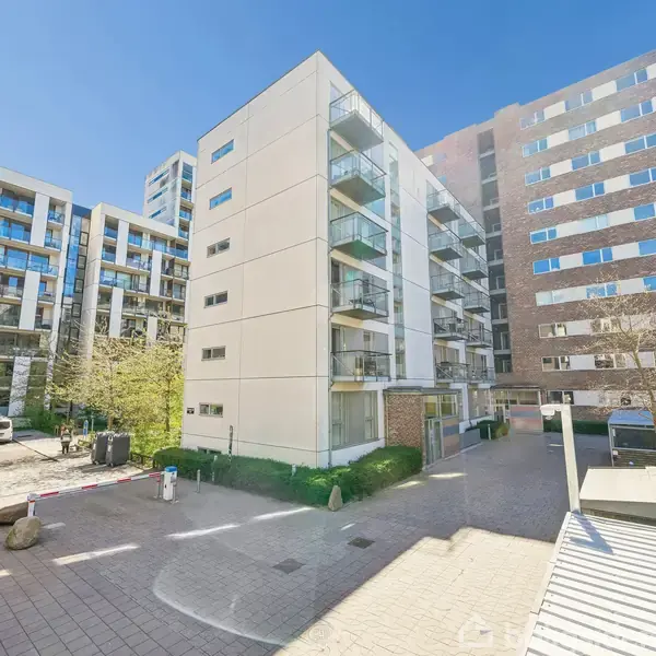 En række moderne lejlighedsbygninger står side om side med glasbalkoner. Solen skinner lyst på den asfalterede gårdsplads, hvor træer og buske omkranser området.