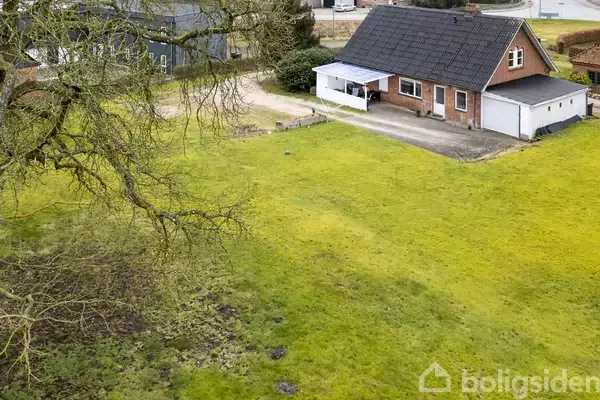 Et enkelt hus med sort tag og rød murstensfacade står stille ved siden af en garage. Haven har en stor, grøn græsplæne, omgivet af træer og nabobygninger.